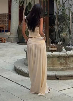Woman in a long beige dress standing in front of a stone fountain in an outdoor setting.