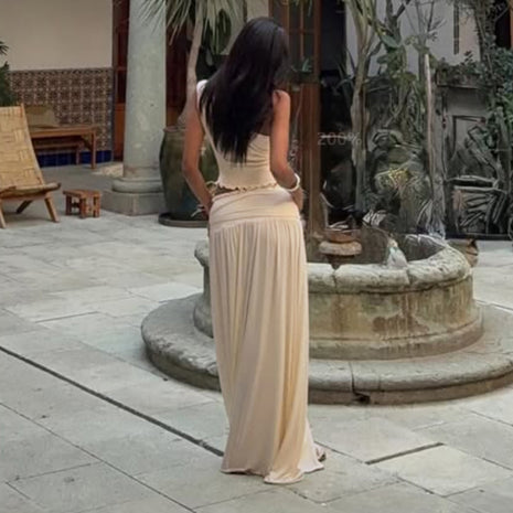 Woman in a long beige dress standing in front of a stone fountain in an outdoor setting.