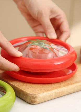 Red silicone lid being placed on a container with a tomato design, on a wooden cutting board.