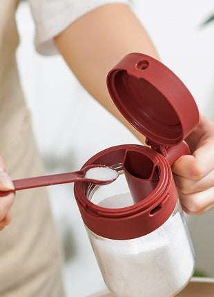 Person using a red measuring spoon to add salt to a clear container.