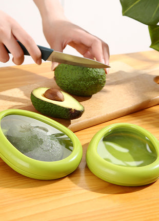 Person cutting an avocado on a wooden board with two green avocado holders in the foreground.